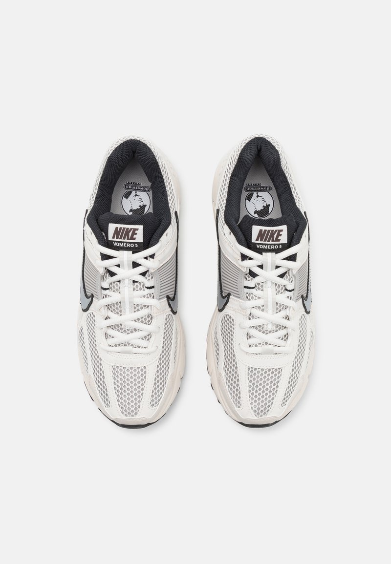 Basket Nike Vomero 5 avec une tige en maille blanche, des accents noirs et une semelle texturée. Comprend des lacets et un logo "Nike" sur la languette.