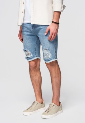 Mand iført lyseblå revnede denimbukser, beige sneakers, hvid t-shirt og lys beige jakke med optrukne ærmer.