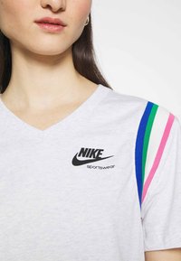  Närbild av en person som bär en ljusgrå Nike sporttröja med V-hals och blå, gröna och rosa ränder på axeln.