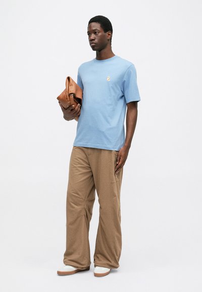 Μπλε κοντομάνικο μπλουζάκι με μικρή γραφική εικόνα, συνδυασμένο με ανοιχτό καφέ παντελόνι σε φαρδιά γραμμή και λευκά παπούτσια, κρατώντας μια καφέ δερμάτινη τσάντα.