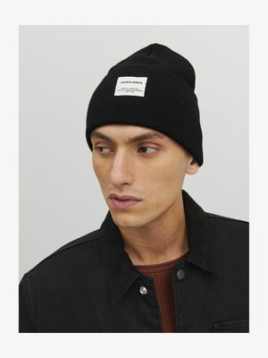 Berretto lavorato a maglia nero con un'etichetta rettangolare bianca con la scritta "JACK & JONES". Indossato da un modello con giacca scura e camicia marrone a trama.