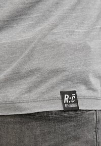 Grå bomulds t-shirt med en let struktureret overflade, der har et sort mærke med "R:C" og "RE:COVERED" tekst langs sømmen.