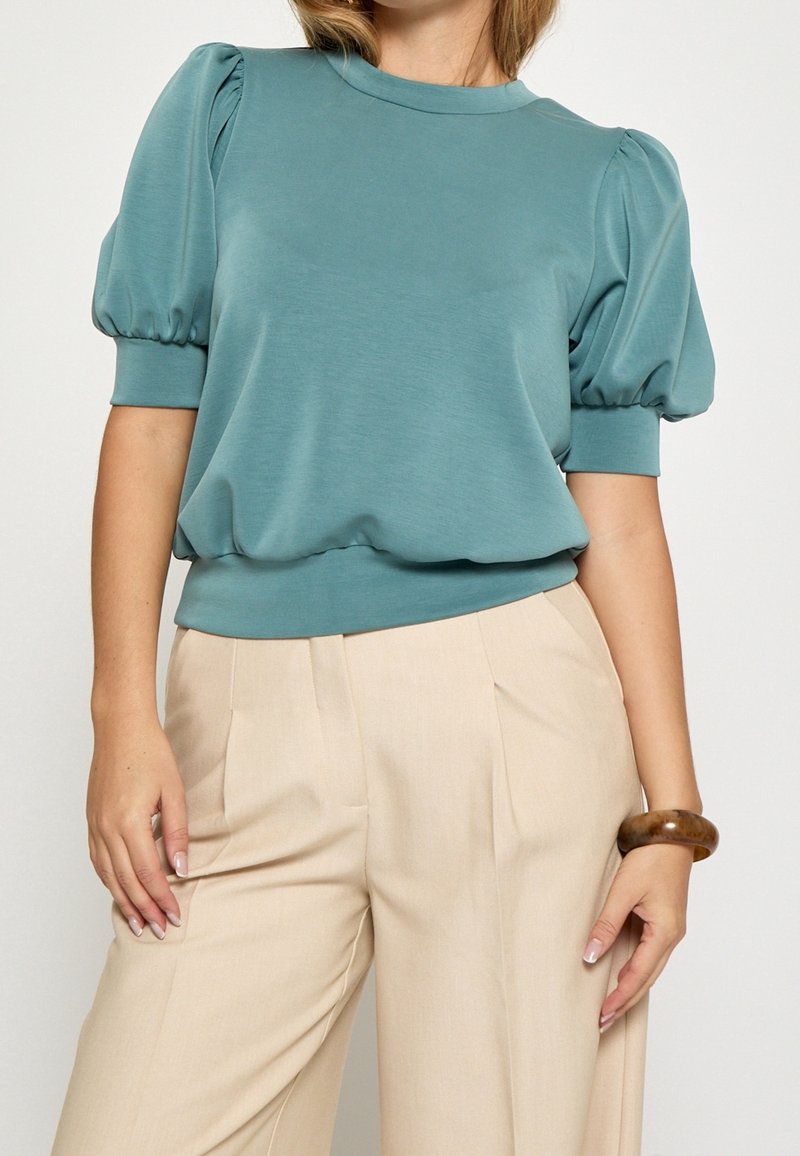 Femme portant un haut bleu sarcelle à manches bouffantes avec des poignets côtelés et un pantalon plissé beige taille haute, accessoirisée d'un bracelet marron au poignet droit.