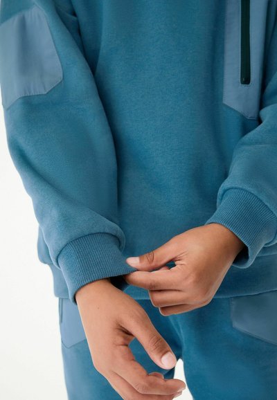 Felpa blu con polsini a coste e dettagli laterali. Il tessuto sembra morbido, con colletto con zip e un motivo testurizzato sulle maniche.