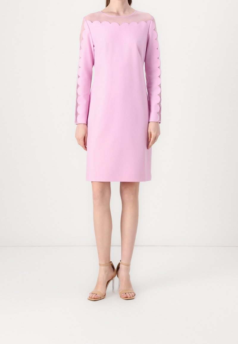 Femme portant une robe rose clair longueur genou avec des bords festonnés et des panneaux transparents sur le haut de la poitrine et les manches, associée à des sandales à talons nude.