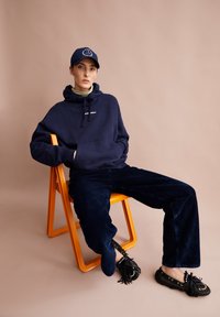 Felpa blu con tasca a marsupio, pantaloni in corduroy blu e scarpe mocassino nere con nappine, seduto su una sedia arancione.