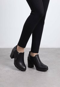 Botas de cuero negro hasta el tobillo con un tacón ancho, paneles laterales elásticos y tachuelas decorativas en la parte trasera. Suela texturizada para un mejor agarre.