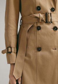 Tan trench coat featuring a double-breasted design, large brown buttons, and a fabric belt with square buckle. Textured cotton material.