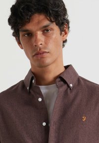 Camicia button-up marrone con tessuto texturizzato, caratterizzata da bottoni bianchi e un logo ricamato arancione sul petto. T-shirt bianca sottostante.
