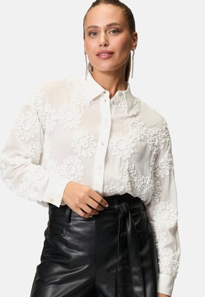 Camicia bianca con ricami floreali testurizzati; tessuto trasparente, maniche lunghe, colletto, abbinata a pantaloni in pelle nera a vita alta.