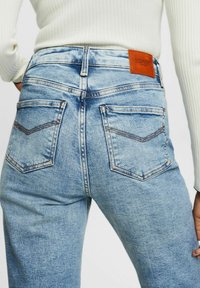 Vaqueros de mezclilla azul claro de talle alto, con un parche de cuero, cinco bolsillos y un sutil detalle de costura en los bolsillos traseros.