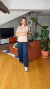 Γυναίκα με μακριά μαλλιά, μπεζ μπλουζάκι και μπλε τζιν στέκεται με σταυρωμένα τα χέρια σε ένα σαλόνι με πορτοκαλί καναπέ και μεγάλο γλαστρόφυτο.