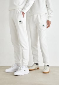 Bílé bavlněné tepláky s elastickými manžetami, se zeleným logem a názvem značky Lacoste. V kombinaci s bílými a béžovými teniskami.