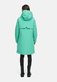 Mintgroene, gevoerde parka met een grote capuchon, voorzien van een uitsnijding aan de achterkant en ritssluiting, gecombineerd met zwarte skinny broeken en laarzen.