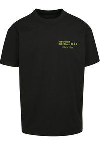 Svart bomulls t-skjorte med løs passform. Har gul tekst foran, inkludert "The Greatest" og detaljer i en grafisk stil.