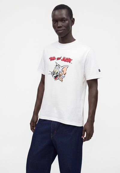 Homme portant un T-shirt blanc avec un graphisme coloré de Tom et Jerry et un pantalon bleu foncé, debout devant un fond uni.