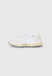Zapatillas Lacoste con una parte superior de malla y cuero blanco, acentos amarillos, suela de goma texturizada y clavos negros para tracción.