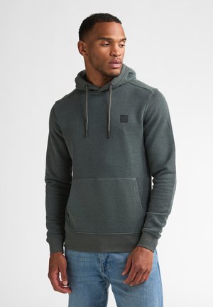 Mann trägt einen dunkelgrünen Kapuzenpullover mit Fronttasche und Kordelzug, kombiniert mit hellblauen Jeans, blickt nach links.