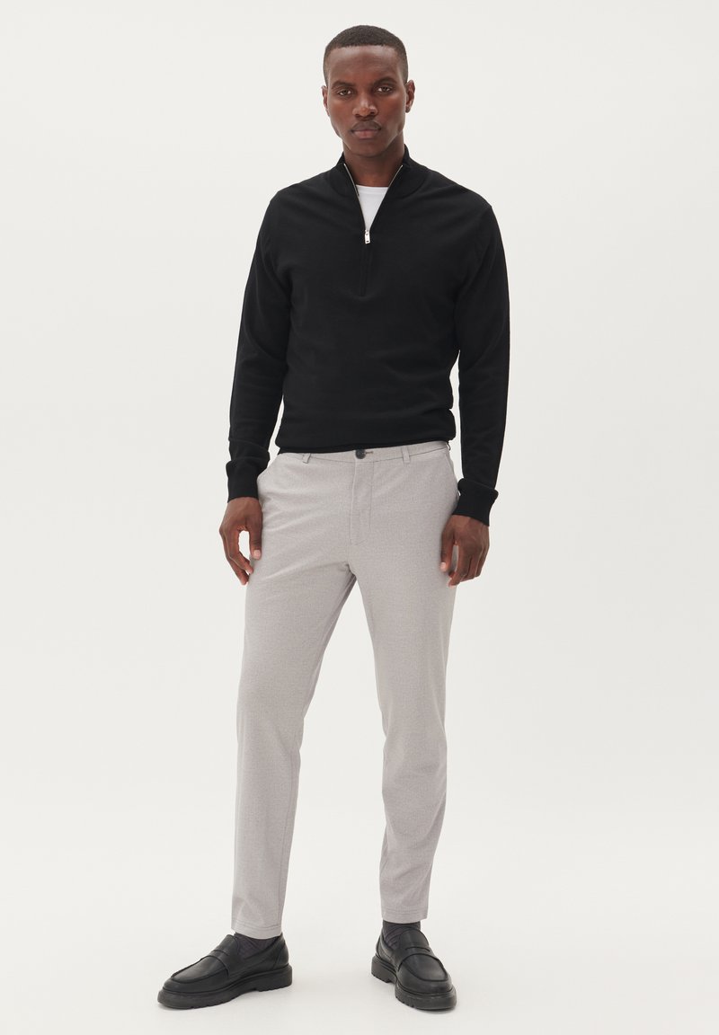 Fekete, cipzáras pulóver hosszú ujjal, világosszürke nadrággal párosítva. A lábbeli sötét, textúrázott felülettel rendelkezik. Egyszerű, modern dizájn.