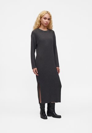 Femme aux longs cheveux blonds bouclés portant une robe midi côtelée à manches longues gris foncé avec une fente latérale et des bottines noires, debout sur fond blanc.