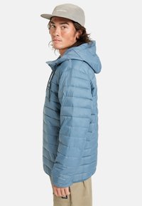 Ljusblå isolerad pufferjacka med huva, med dragkedja fram, horisontell quiltning och logodetalj på bröstet.