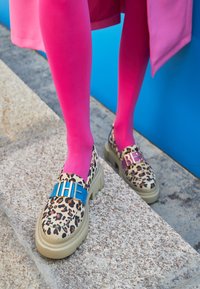 Le scarpe con suola piatta a stampa leopardo presentano un dettaglio clip blu e rosa sulla parte anteriore, abbinate a collant fucsia su uno sfondo blu.