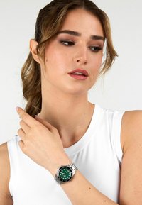 Orologio in acciaio inossidabile argentato con quadrante verde, indicatori delle ore bianchi e bracciale metallico indossato su un polso. Presenta una cassa e una lunetta rotonde.