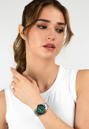 Orologio in acciaio inossidabile argentato con quadrante verde, indicatori delle ore bianchi e bracciale metallico indossato su un polso. Presenta una cassa e una lunetta rotonde.