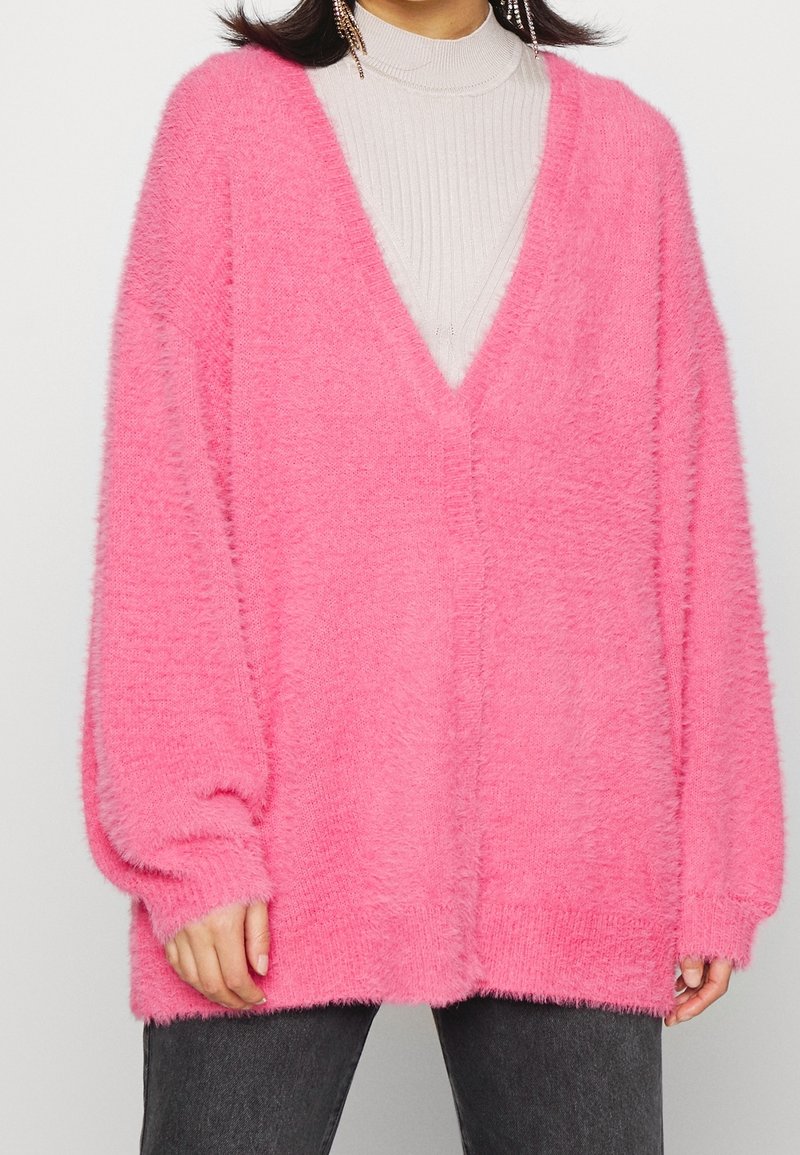 Fluffy pink oversized sweater with a deep V-neck, long sleeves, and ribbed texture. Soft material contrasts with the smooth inner layer.