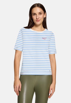 Kvinde iført en lyseblå og hvidstribet kortærmet skjorte med "merci" og et lille hjerte broderet på brystet, kombineret med olivengrønne bukser.