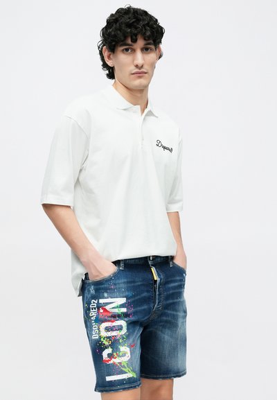 Polo blanc à manches courtes avec un petit logo noir ; associé à un short en denim bleu avec des éclaboussures de peinture colorées et du texte.