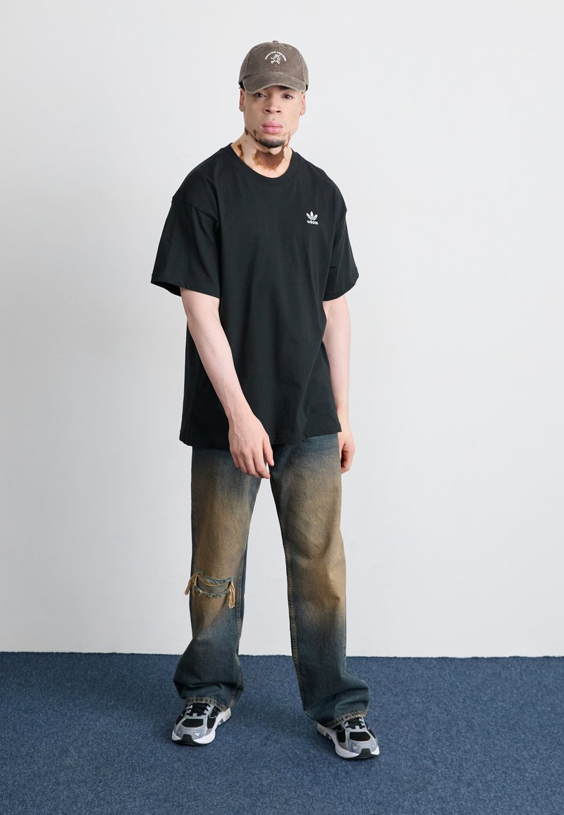 Schwarzes Baumwoll-T-Shirt mit kurzen Ärmeln, hellblaue bis beige Jeans mit einem aufgerissenen Knie und graue Mütze. Details umfassen ein Logo auf dem Shirt.