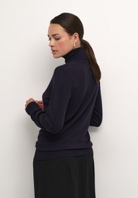 Marineblauwe coltrui gemaakt van zachte stof, slim-fit ontwerp, geribde manchetten en zoom, gecombineerd met een zwarte rok die een gladde textuur toont.