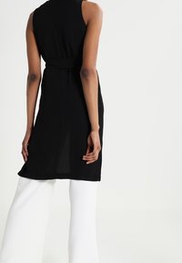 Schwarzes ärmeloses Kleid mit einer taillierten Oberseite und einem taillierten Gürtel, kombiniert mit weiten weißen Hosen. Glattes Material und klare Linien.