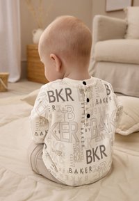 Body à manches longues en tissu blanc avec motifs de texte gris, doté d'un col rond et de trois boutons noirs au dos. Pantalon gris assorti en dessous.