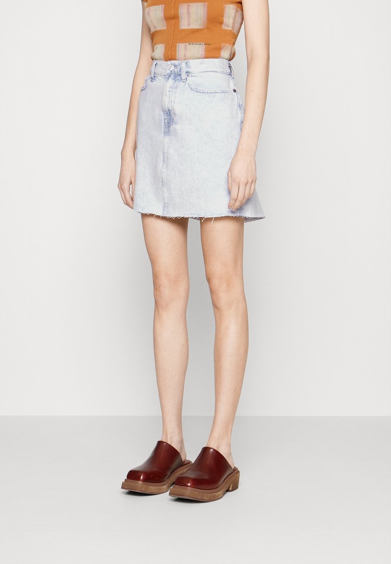 Personne portant une mini-jupe en jean bleu clair, un haut sans manches orange et des sabots en cuir marron, debout devant un fond uni.