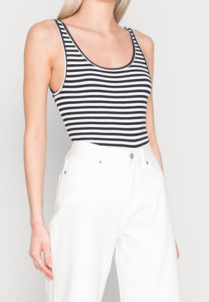 Frau trägt ein ärmelloses schwarz-weiß gestreiftes Oberteil mit horizontalen Streifen, das in eine hoch taillierte weiße Hose gesteckt ist, vor einfachem Hintergrund.