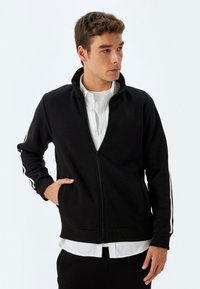 Chaqueta negra con cremallera en tela suave, que presenta un cuello alto, franjas laterales blancas, puños acanalados y un bolsillo con cremallera en el frente.