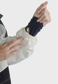 Un poignet de veste blanc cassé avec des fronces élastiques présente une manche en polaire bleu marine en dessous et une étiquette marron avec un point blanc.