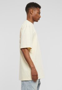 Cream-colored cotton t-shirt, relaxed fit, short sleeves, featuring a graphic design on the back. Worn with light blue denim jeans.