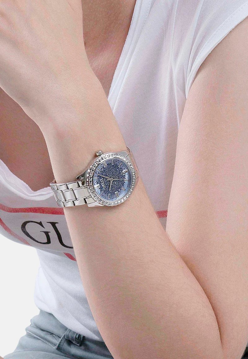 Srebrny zegarek z błyszczącą niebieską tarczą, kryształowymi akcentami wokół obręczy i metalową bransoletą połączoną noszoną na nadgarstku.
