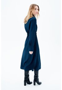 Frau mit langen blonden Haaren, die ein navyblaues langärmliges Kleid und schwarze kniehohe Stiefel mit Absatz trägt, steht vor einem weißen Hintergrund.