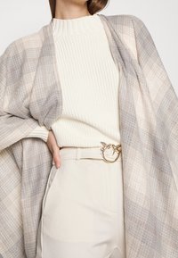 Donna che indossa un maglione a coste color crema infilato in pantaloni beige a vita alta con una cintura a fibbia dorata a forma di uccello e un leggero scialle a quadri sulle spalle.