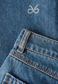 Keskmise sinise tooniga teksakangas lähivõttega taskust. Kattel on väikesestest kristallidest valmistatud logo, mis rõhutab kanga tekstuuri.