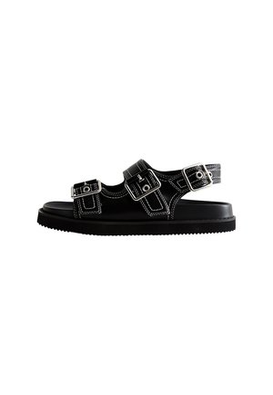 Sort sandal med åben tå med tre justerbare sølvspænder og hvide syninger på en flad gummisål, vist fra siden på hvid baggrund.