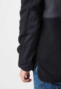 Giacca in fleece nera con materiale testurizzato. Presenta un tessuto liscio sulle maniche, un pannello grigio a contrasto e lati con zip.