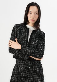 Black and white tweed jacket with a textured finish, featuring a round neckline, no buttons, and an accent flower detail on the left side.