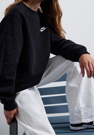Sweatshirt noir avec un logo Nike blanc sur la poitrine, associé à un pantalon large blanc et des baskets noires, assis sur un banc bleu.