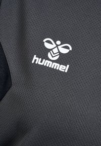 Hvid styliseret bi-logo med teksten "hummel" på mørkegrå tekstureret, åndbar stof, sandsynligvis del af sportstøj eller atletisk tøj.