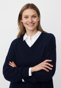 Granatowy sweter z dekoltem w kształcie litery V i prążkowanymi detalami, noszony na białej koszuli z kołnierzykiem. Miękka tekstura, długie rękawy, skrzyżowane ramiona.